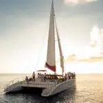A catamaran sits on the ocean during a Red Sail Sports' Sunset Sail in Grand Cayman. We view it from behind, the front of the boat facing toward the horizon. Numerous people stand around on the deck, enjoying the atmosphere. A white and red sail soars above them. Another catamaran is visible in the distance, the sky above both is a pale blue and white as the sun shines bright.