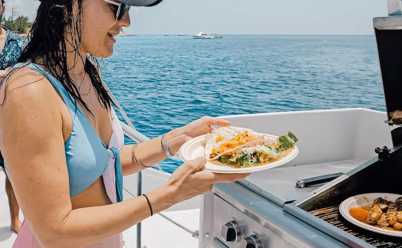 A lady in swimwear, a baseball cap, and sunglasses holds a plate while standing in front of a partially visible BBQ grill. A taco and salad are already on her plate. The lady’s hair is wet, indicating that she has been enjoying some watersports in Grand Cayman. The blue ocean and clear sky are visible in the background. A lady in swimwear, a baseball cap, and sunglasses holds a plate while standing in front of a partially visible BBQ grill. A taco and salad are already on her plate. The lady's hair is wet, indicating that she has been enjoying some watersports in Grand Cayman. The blue ocean and clear sky are visible in the background.