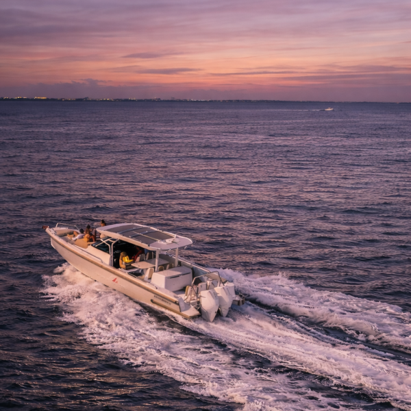 An aerial shot of a Red Sail Sports 37’ Axopar boat speeding through the ocean water surrounding Grand Cayman. We can see some people relaxing on the front section, with a few more sitting under the roof covering in the vessel’s midsection. It's twilight, so the sky is darkening and a dull orange at the horizon.