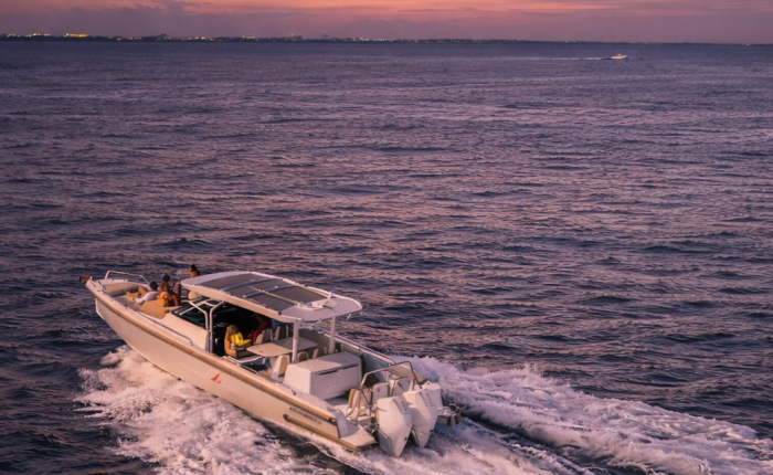 An aerial shot of a Red Sail Sports 37’ Axopar boat speeding through the ocean water surrounding Grand Cayman. We can see some people relaxing on the front section, with a few more sitting under the roof covering in the vessel’s midsection. It's twilight, so the sky is darkening and a dull orange at the horizon.